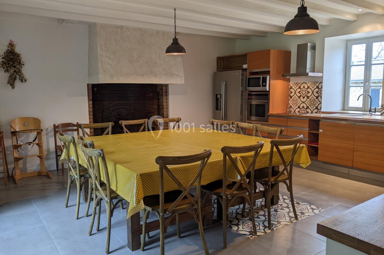 Salle à manger lumineuse avec une grande table recouverte d'une nappe jaune, entourée de chaises en bois, près d'une cuisine…