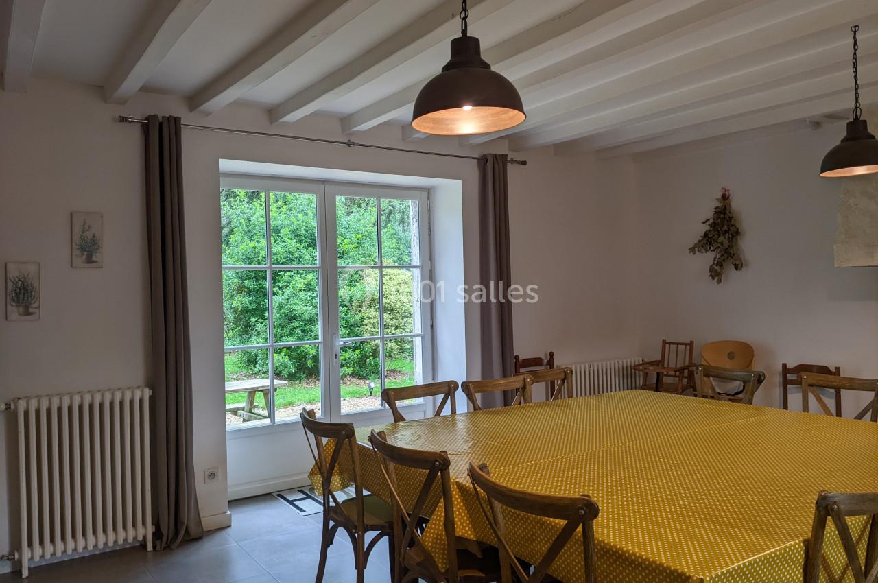 Salle à manger lumineuse avec grande table recouverte d'une nappe jaune, chaises en bois et vue sur un jardin verdoyant.