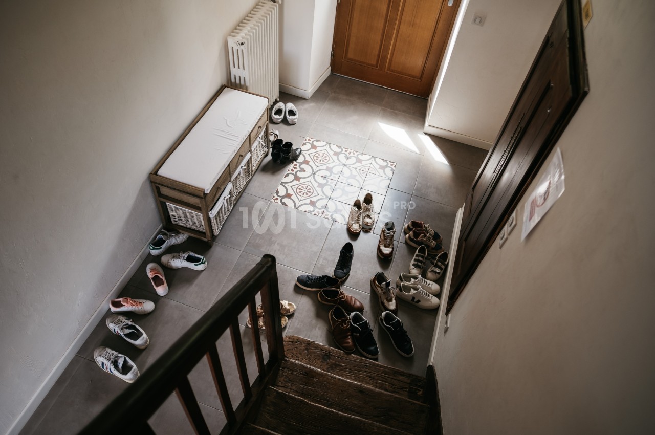 Vue d'une entrée de maison avec plusieurs paires de chaussures dispersées sur le sol carrelé près d'un banc de rangement.