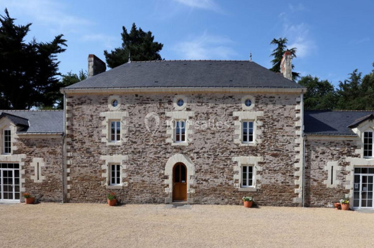 Façade d'une maison en pierre avec toit en ardoise, entourée d'arbres et donnant sur une cour en gravier.