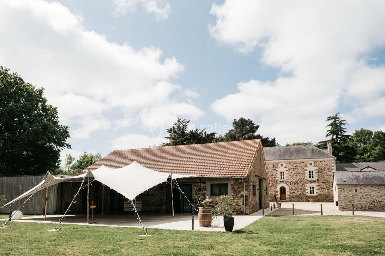 Bâtiment en pierre avec toit en tuiles, tente blanche installée devant, entouré de pelouse et d'arbres.