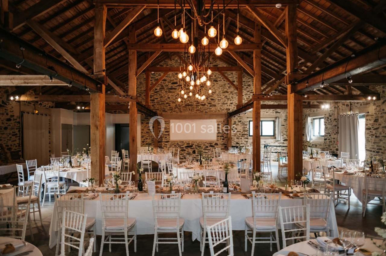 Salle de réception rustique avec poutres apparentes, tables dressées et chaises blanches, éclairée par des suspensions.