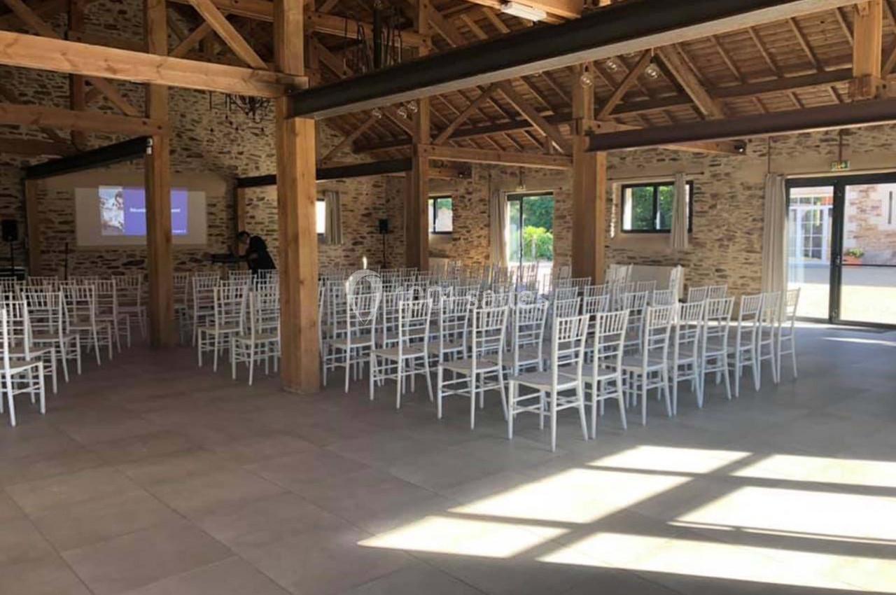 Salle de réception lumineuse avec des chaises blanches alignées et un écran de projection en arrière-plan.