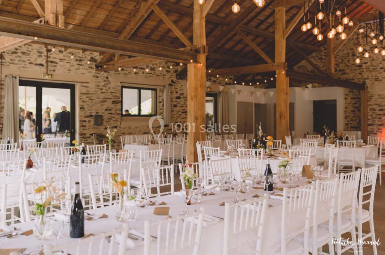 Salle de réception lumineuse avec tables dressées, chaises blanches et décoration florale, sous un plafond en bois.