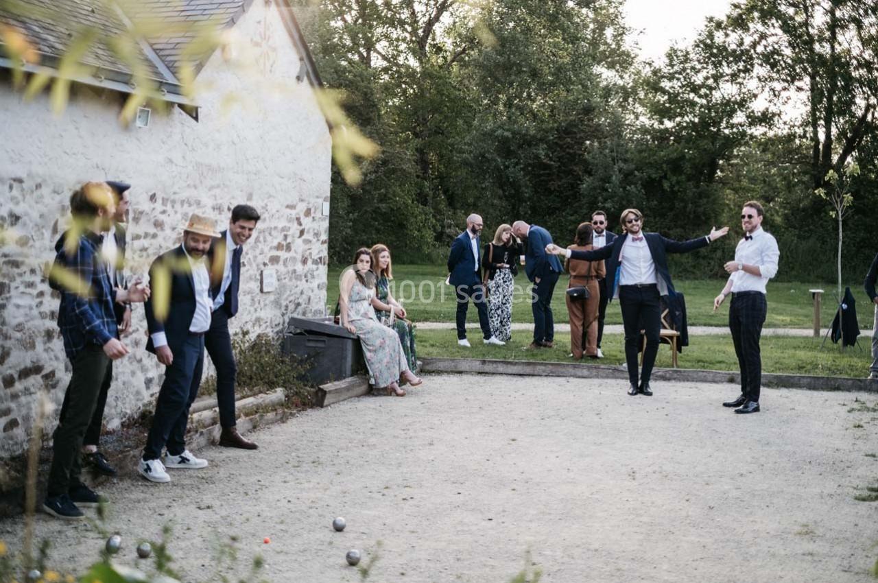 Des personnes jouent à la pétanque dans un jardin, tandis que d'autres discutent près d'une maison en pierre.