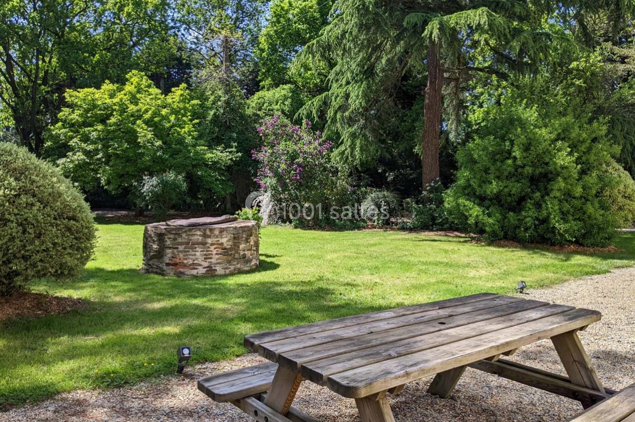 Table de pique-nique en bois sur un sol gravillonné, devant un puits en pierre et un jardin verdoyant avec arbres et…