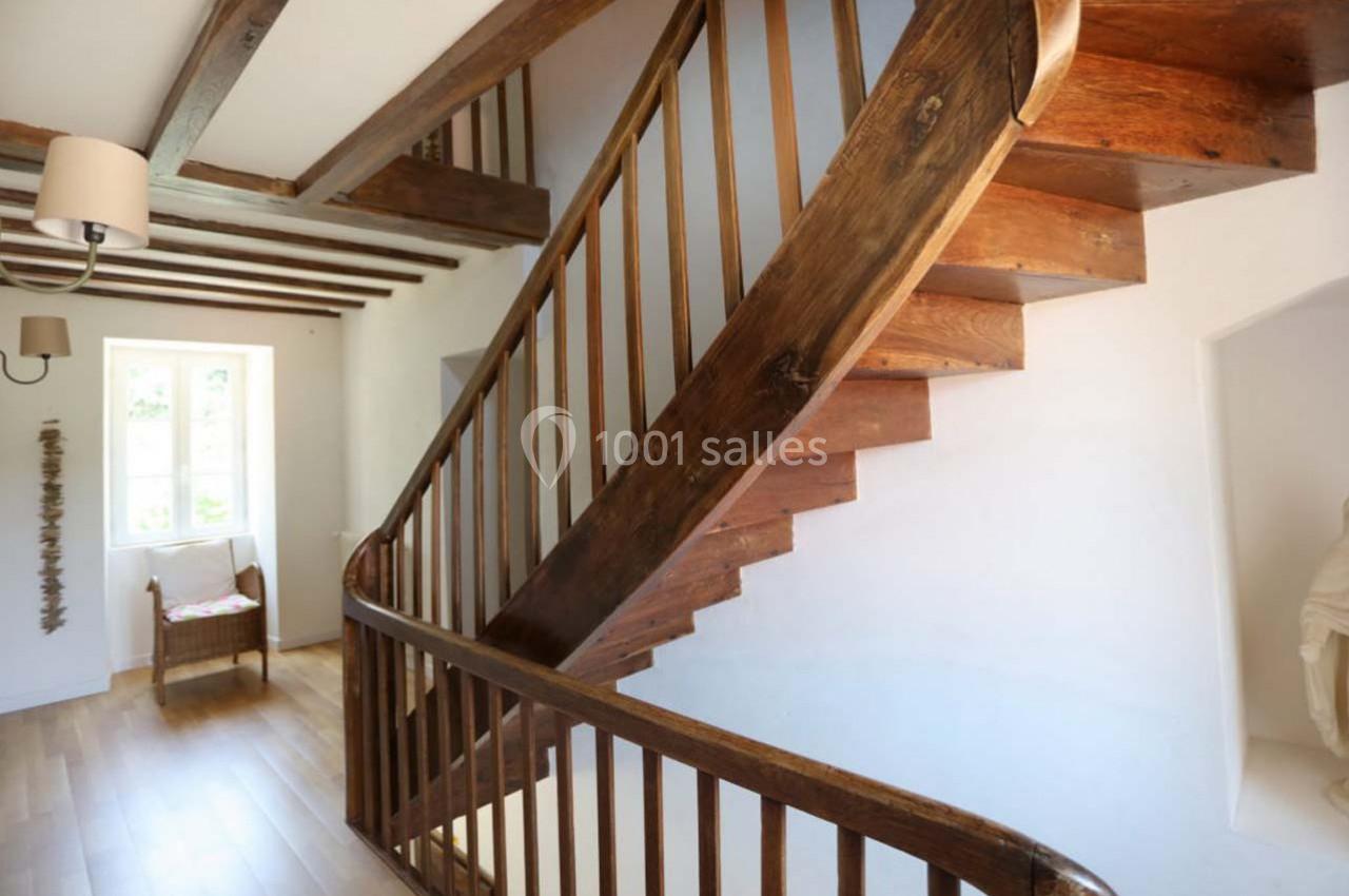 Escalier en bois avec rampe, dans une maison lumineuse au sol en parquet et poutres apparentes au plafond.