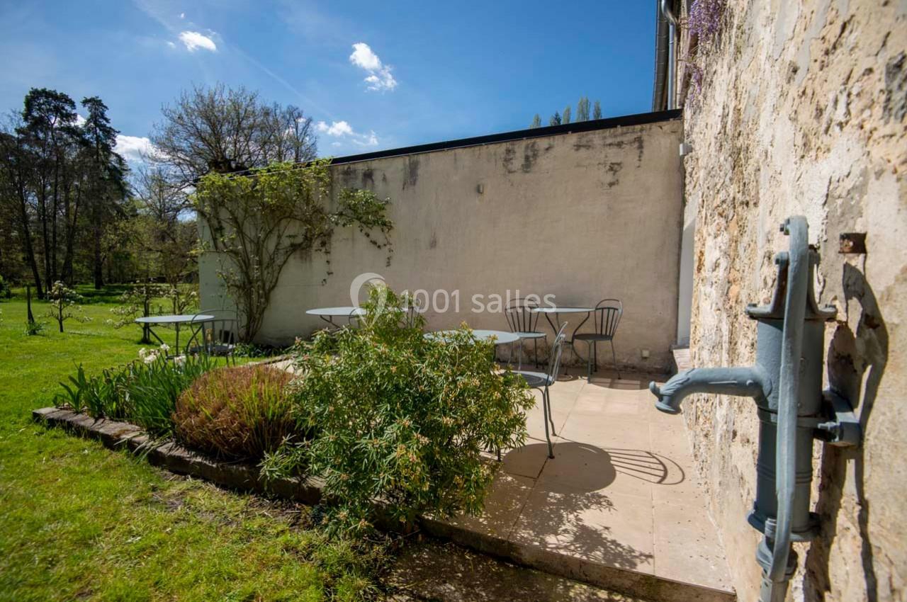 Terrasse extérieure avec tables et chaises en métal, entourée de verdure et adossée à un mur en pierre.