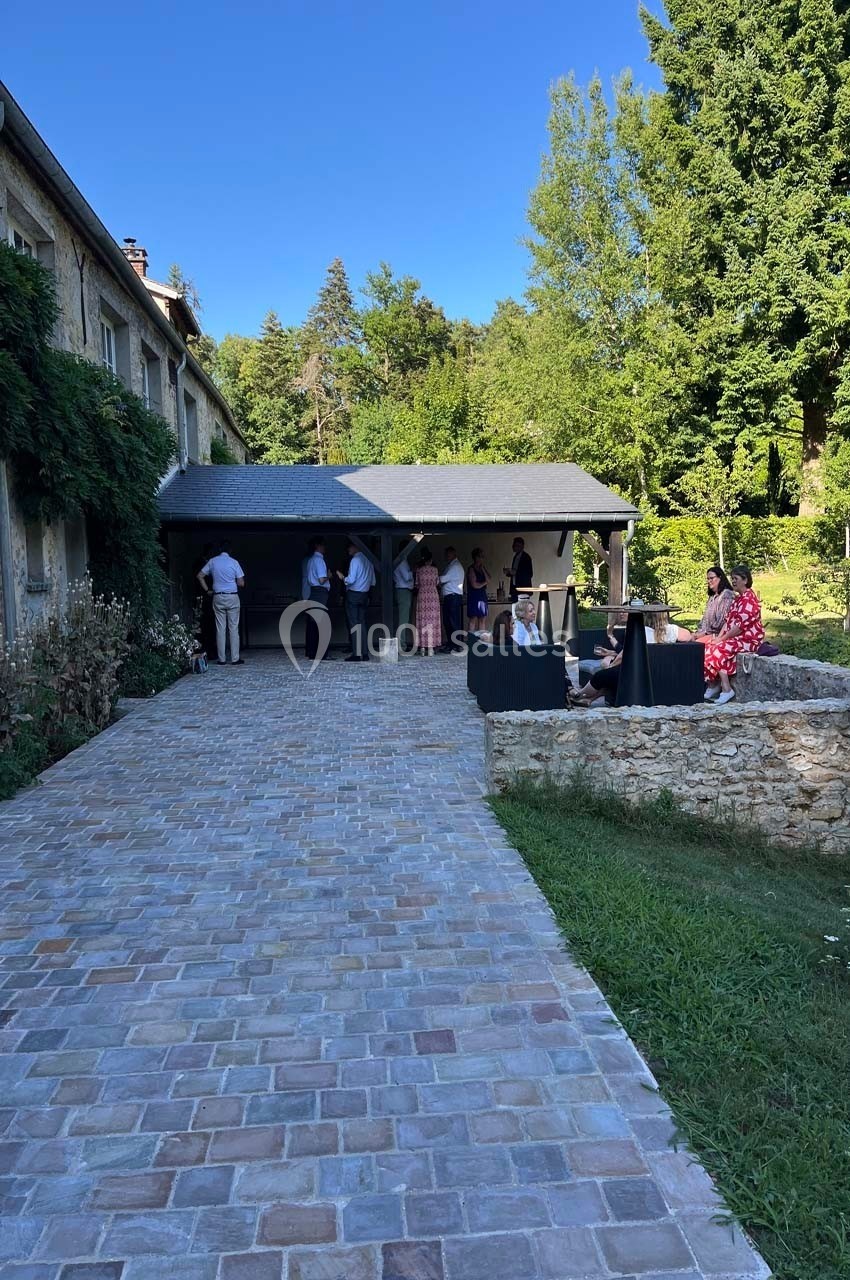 Personnes rassemblées près d'une terrasse en pierre, entourée de verdure et d'un bâtiment en pierre sous un ciel dégagé.
