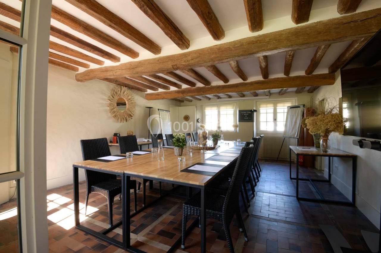 Salle de réunion lumineuse avec table en bois, chaises noires, poutres apparentes et décoration naturelle.