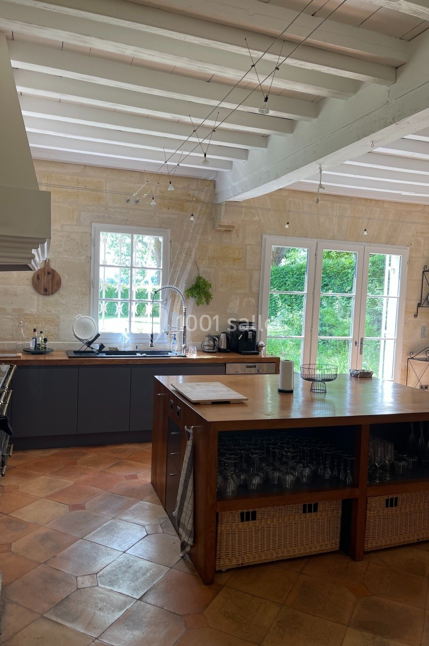 Cuisine lumineuse avec murs en pierre, poutres apparentes, îlot central en bois et grandes fenêtres donnant sur un jardin.