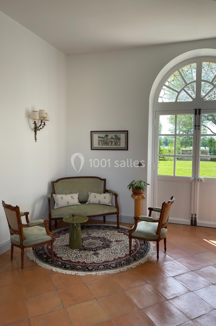 Petit salon lumineux avec canapé, fauteuils anciens, table basse, tapis rond, et vue sur un jardin par une porte-fenêtre.