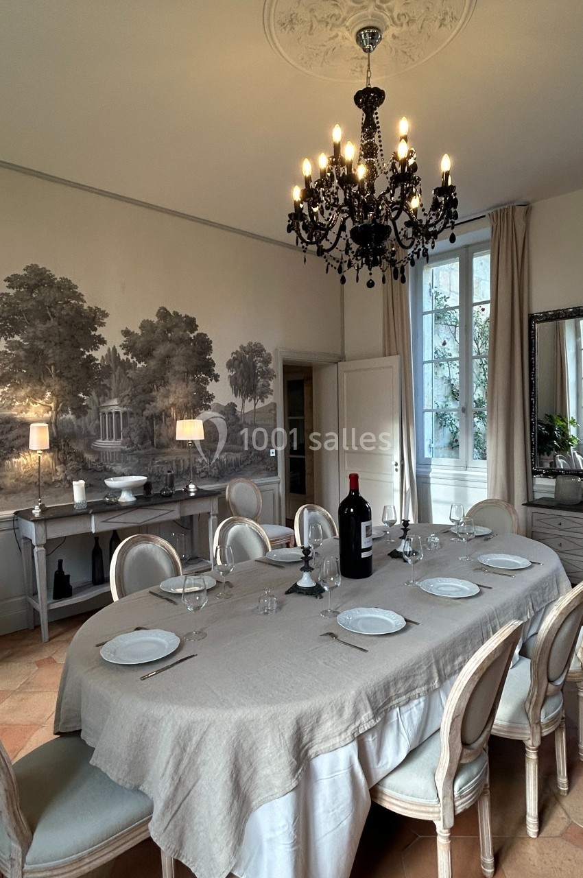 Salle à manger élégante avec une grande table dressée, un lustre noir et une fresque murale représentant un paysage.