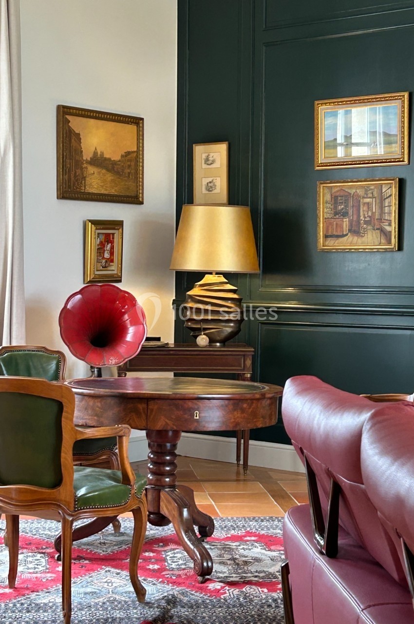 Salon élégant avec table en bois, fauteuils en cuir, gramophone rouge, lampe dorée et tableaux sur un mur vert foncé.