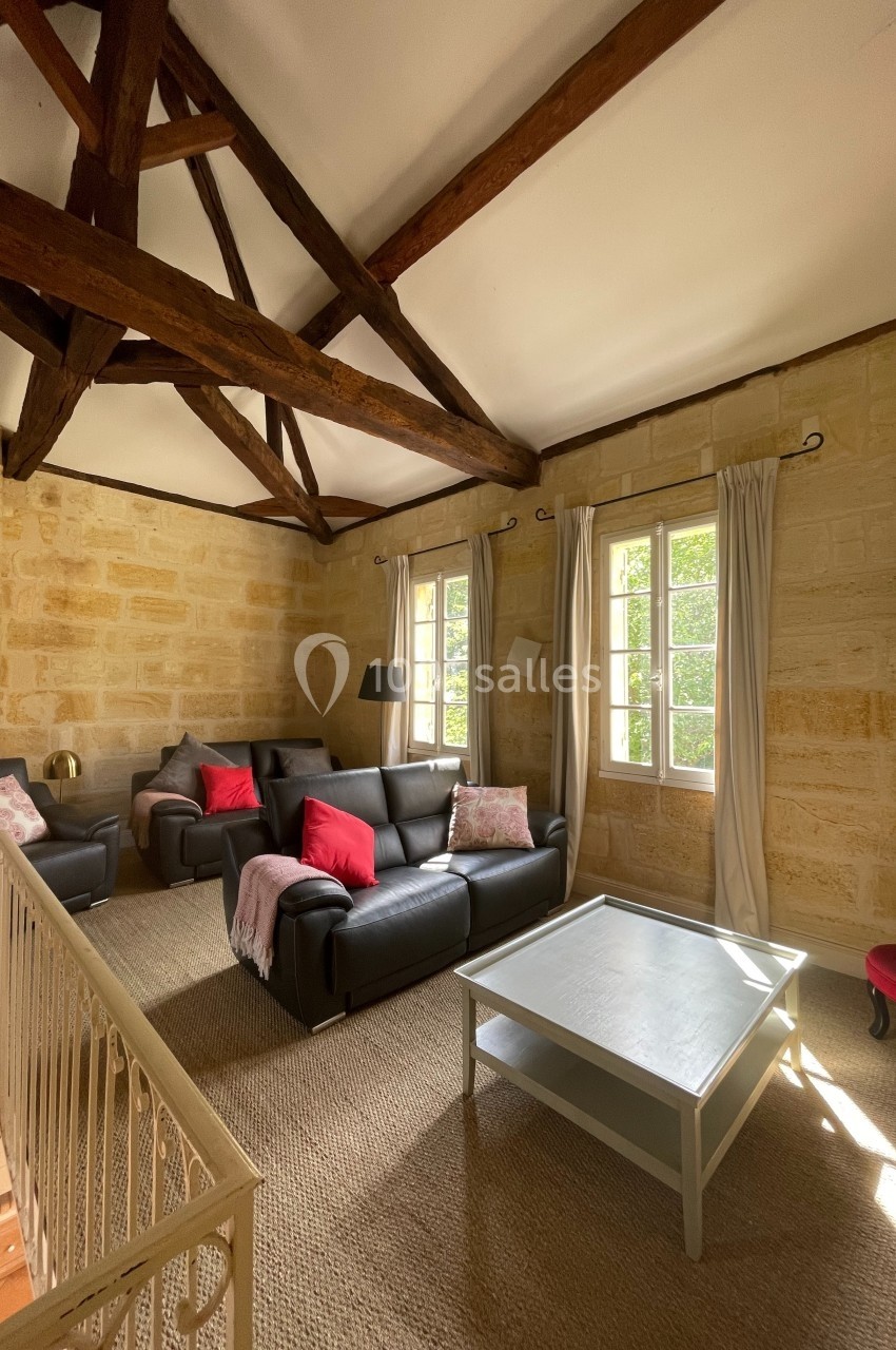 Salon lumineux avec murs en pierre, poutres apparentes, canapés en cuir, table basse et grandes fenêtres.