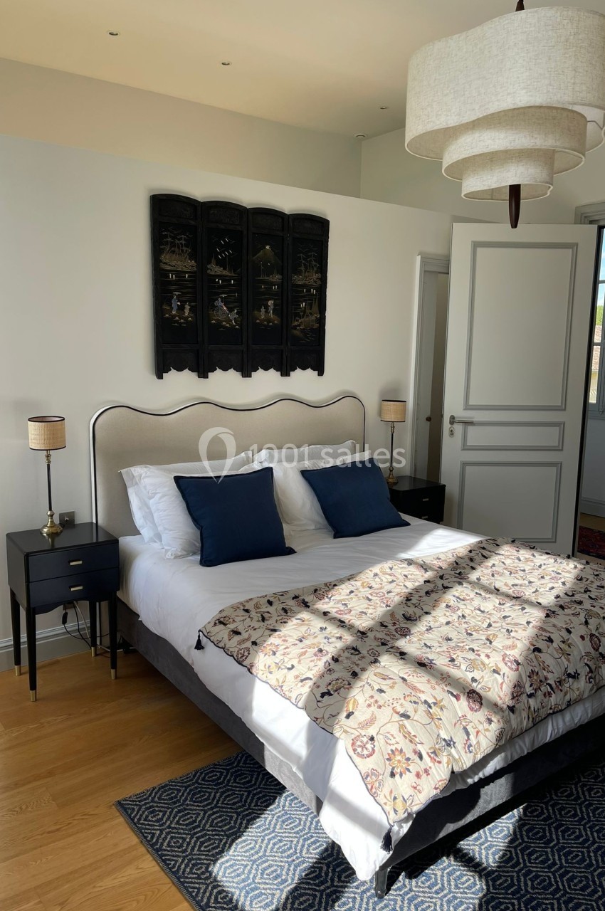 Chambre lumineuse avec lit double, coussins bleus, dessus-de-lit fleuri, lampes de chevet et décoration murale sombre.