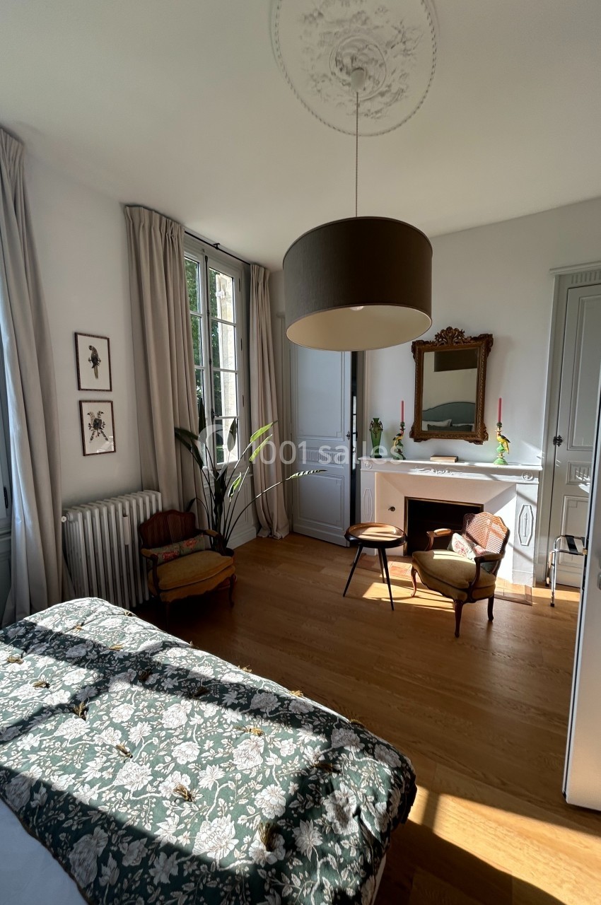 Chambre lumineuse avec lit à motif floral, fauteuils près d'une cheminée, miroir et grandes fenêtres donnant sur l'extérieur.