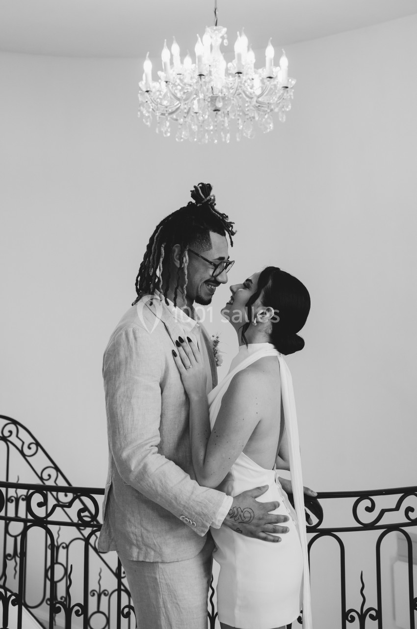 Un couple souriant s'enlace devant une rambarde en fer forgé, sous un lustre élégant, dans un cadre lumineux.