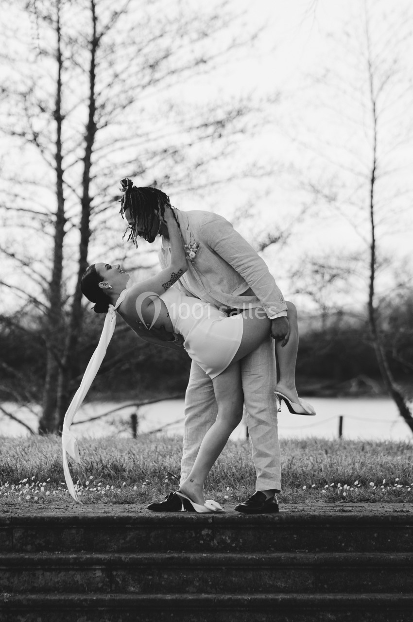 Un couple pose en extérieur, l'un portant l'autre dans une posture romantique, devant un paysage arboré.