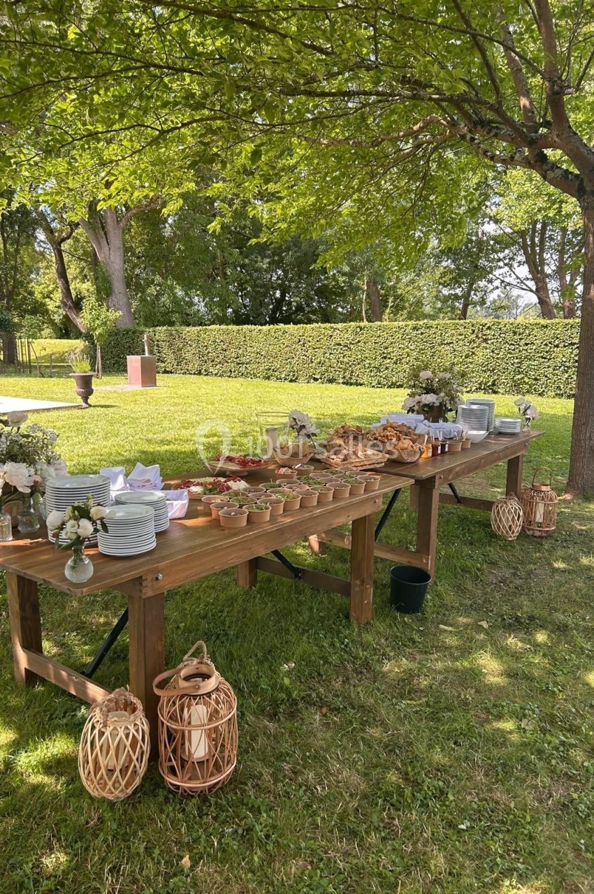Table en bois dressée en extérieur avec vaisselle, plats variés, fleurs et lanternes sous des arbres.