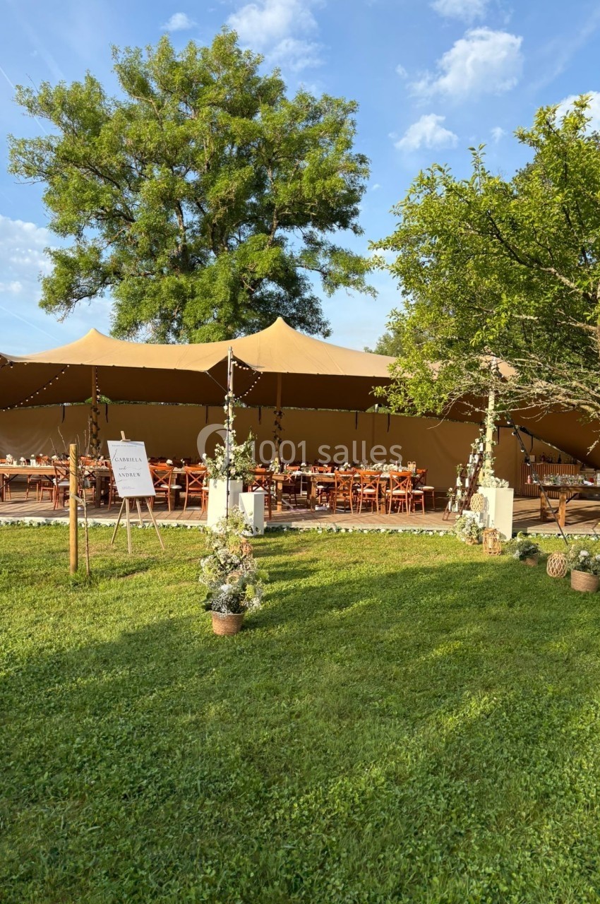 Grande tente beige installée dans un jardin verdoyant, aménagée avec des tables en bois pour un événement en plein air.