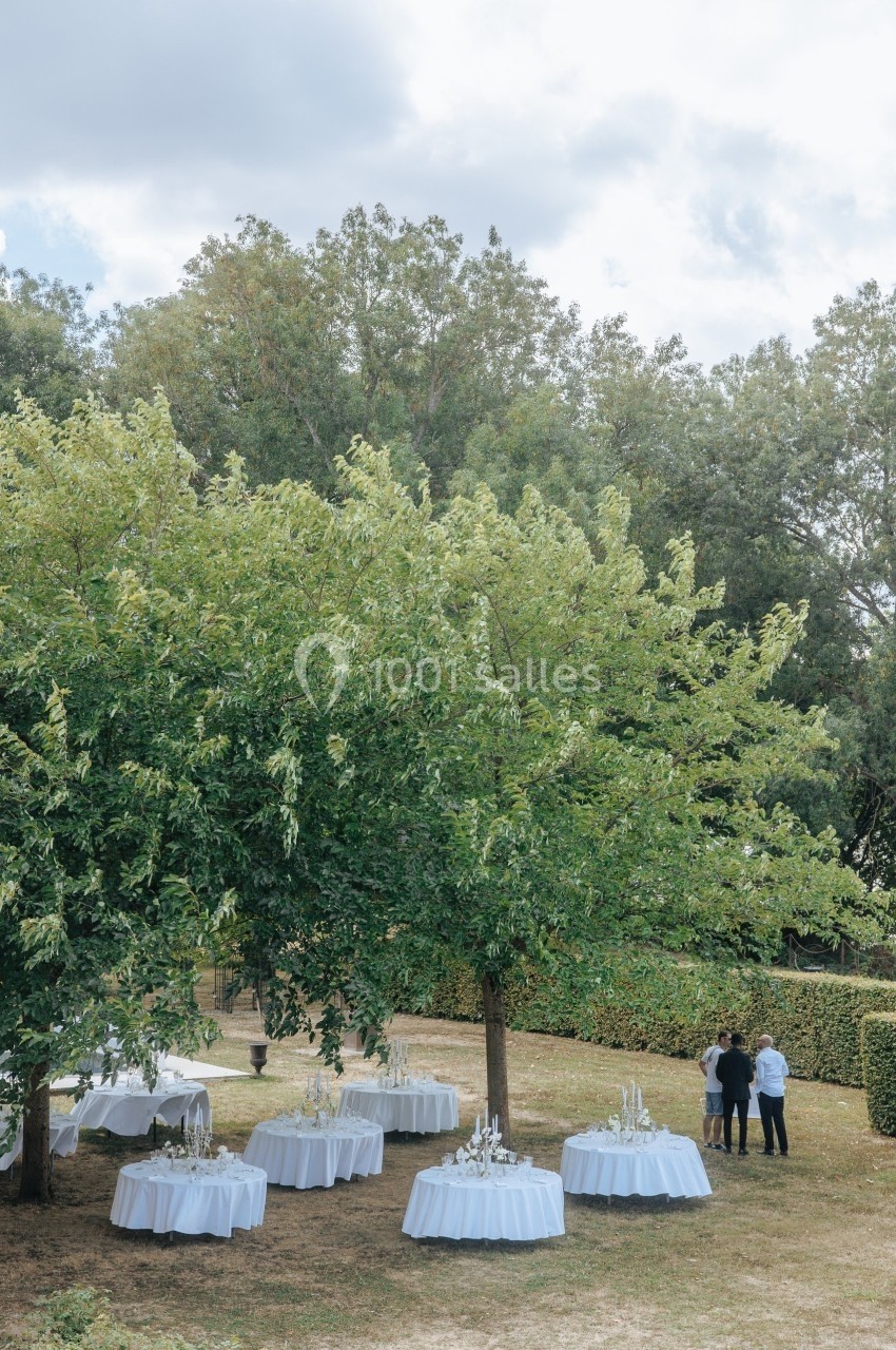 Tables rondes dressées avec nappes blanches sous des arbres dans un jardin, avec trois personnes en arrière-plan.