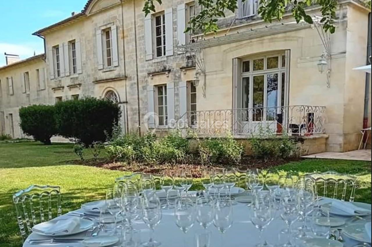 Table dressée avec des verres et assiettes dans un jardin devant une grande maison en pierre avec volets blancs.