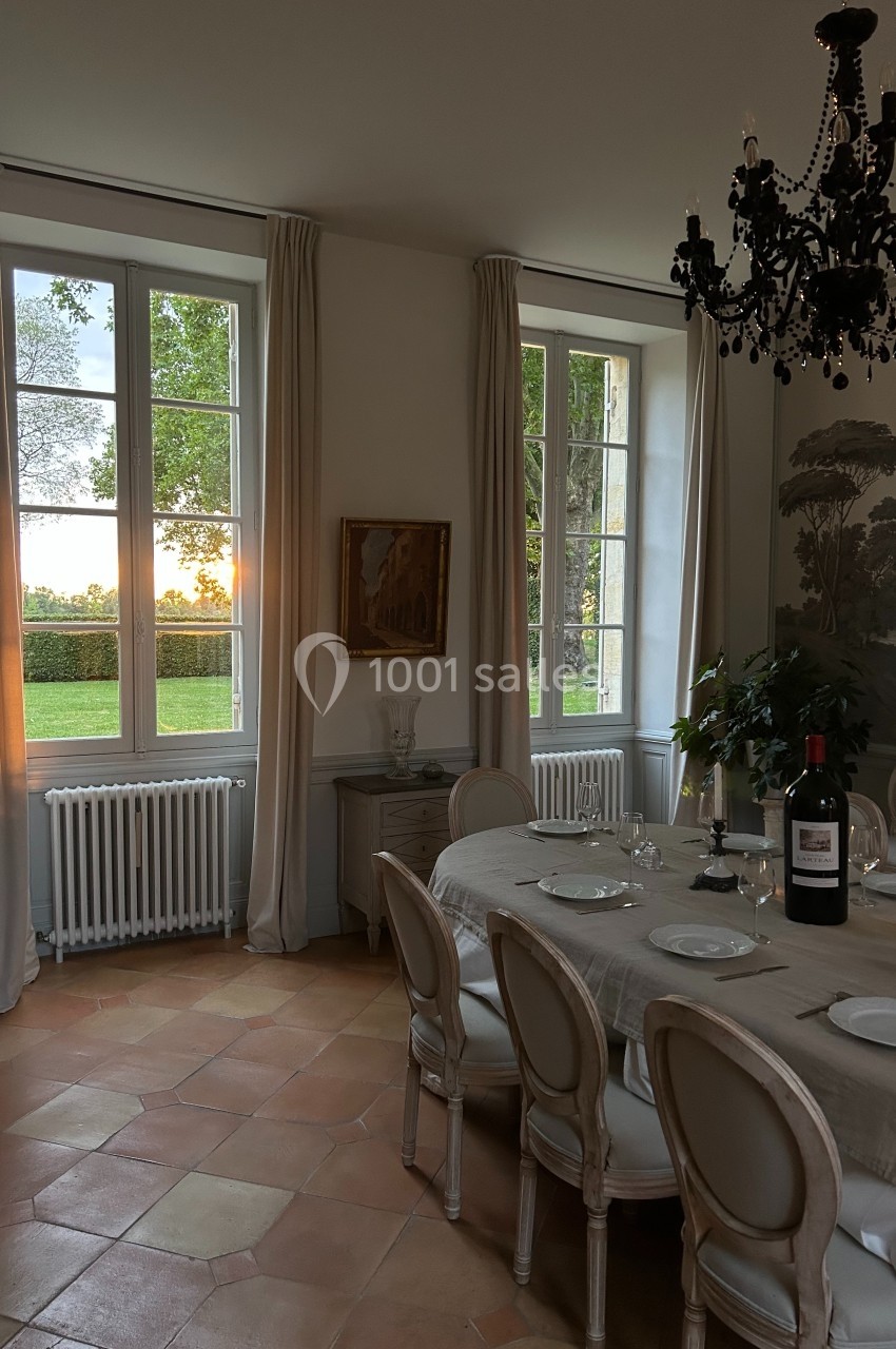 Salle à manger élégante avec table dressée, chandelier noir, grandes fenêtres et vue sur un coucher de soleil.
