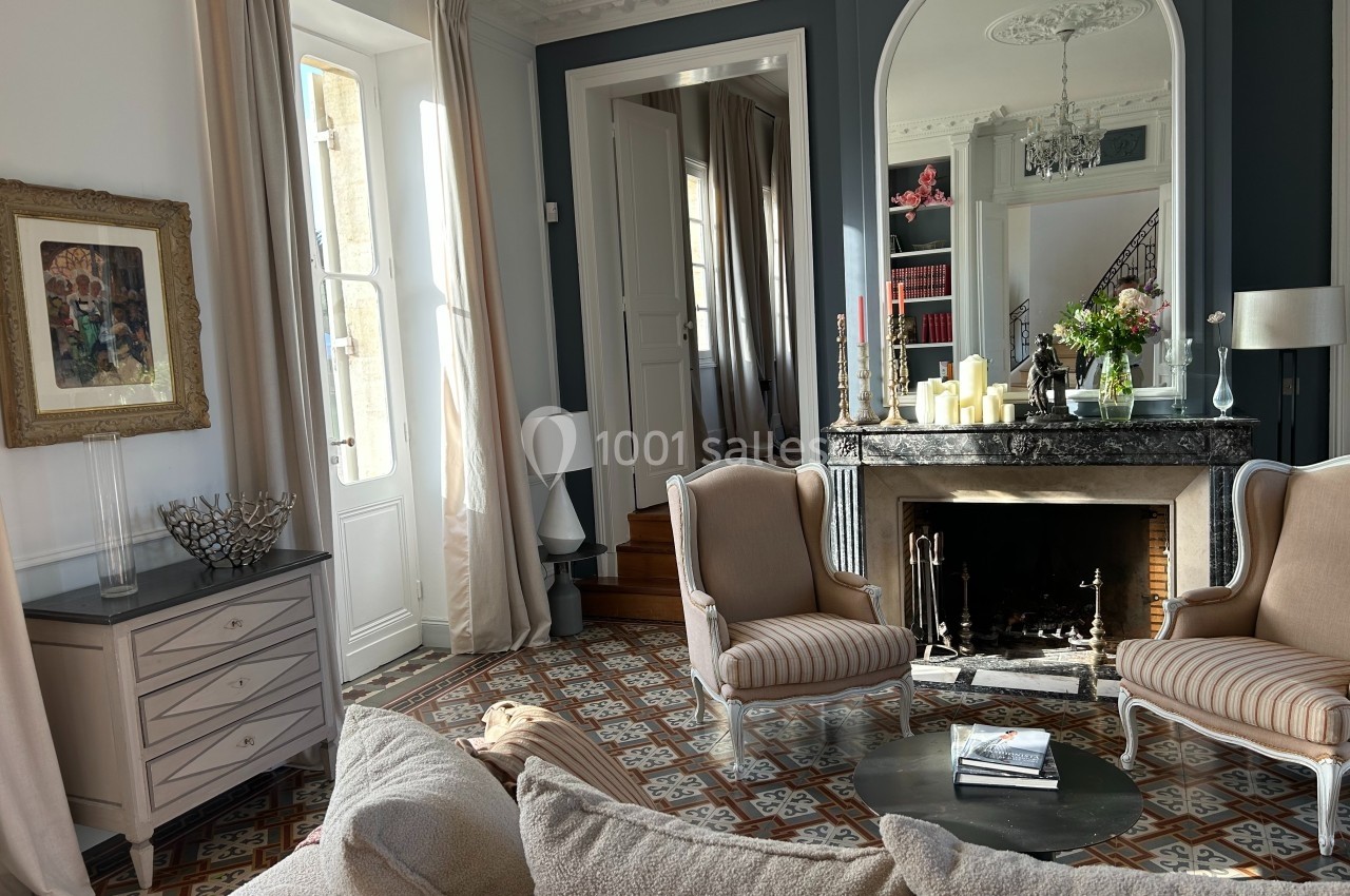 Salon élégant avec cheminée, fauteuils classiques, table basse, grandes fenêtres et carrelage décoratif au sol.