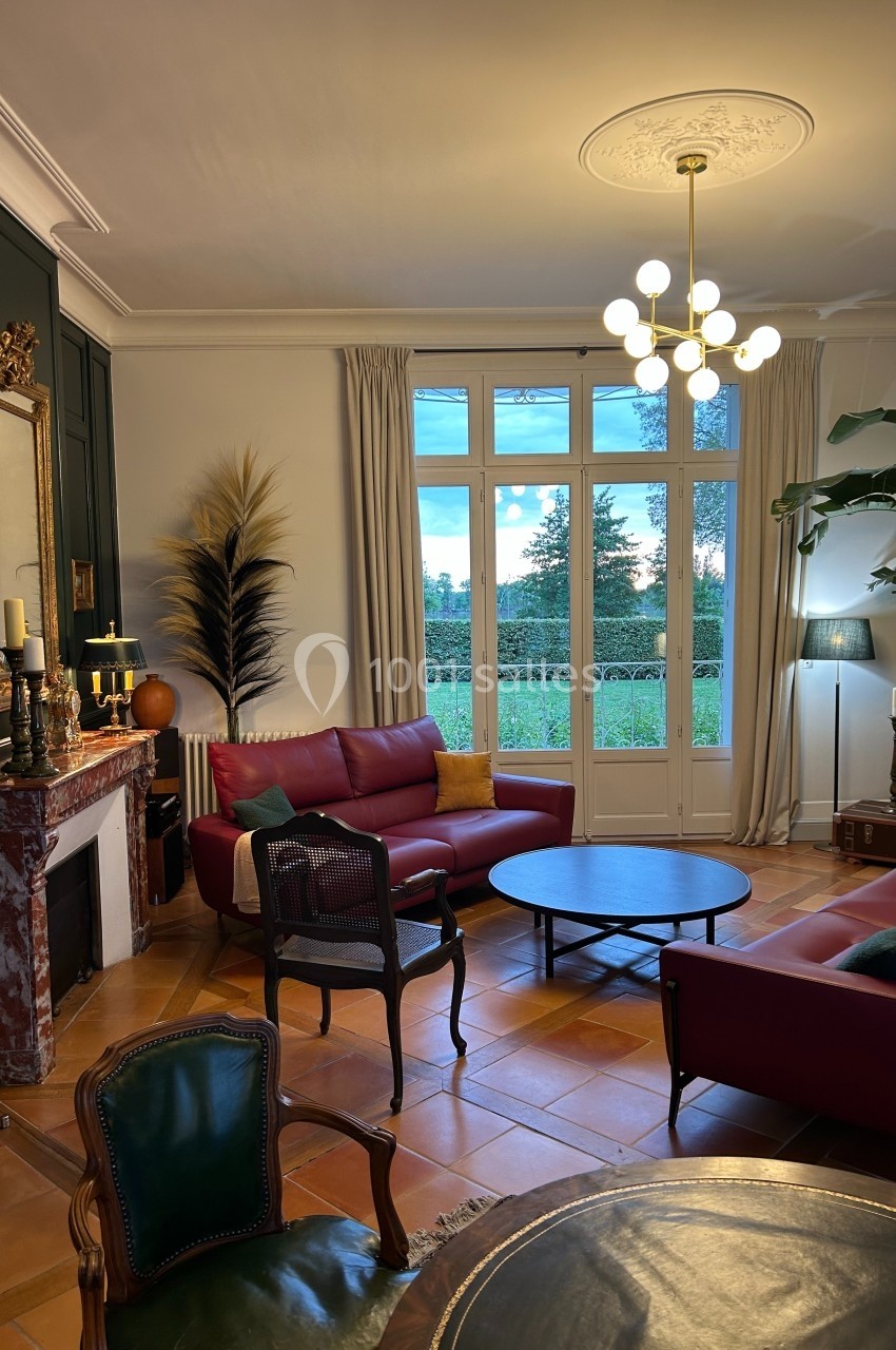 Salon élégant avec canapés rouges, table basse ronde, cheminée en marbre, grandes fenêtres et vue sur jardin.