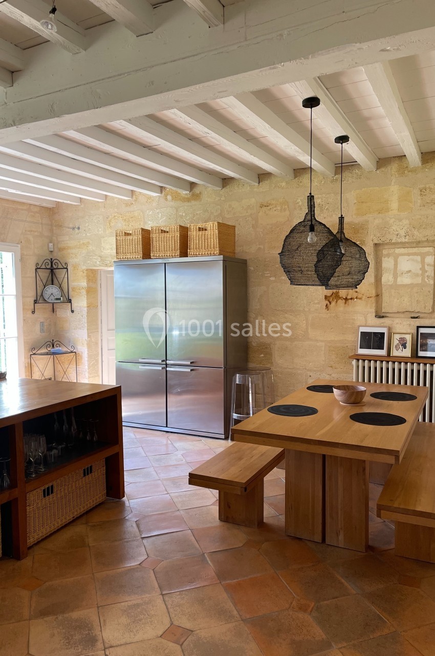 Cuisine lumineuse avec table en bois, bancs assortis, réfrigérateur inox, murs en pierre et suspensions noires.