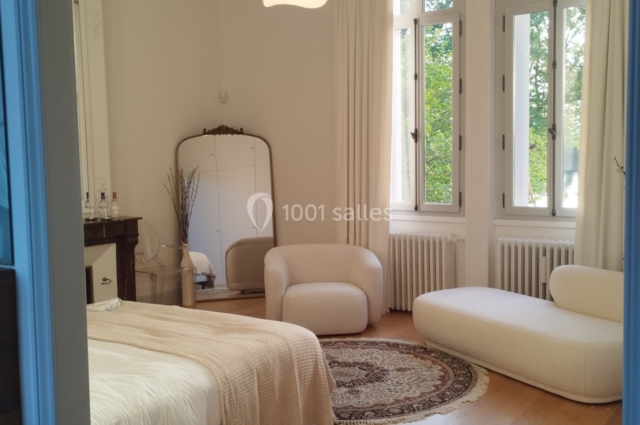 Chambre lumineuse avec lit, fauteuils blancs, miroir ancien, tapis rond et grandes fenêtres donnant sur un espace vert.