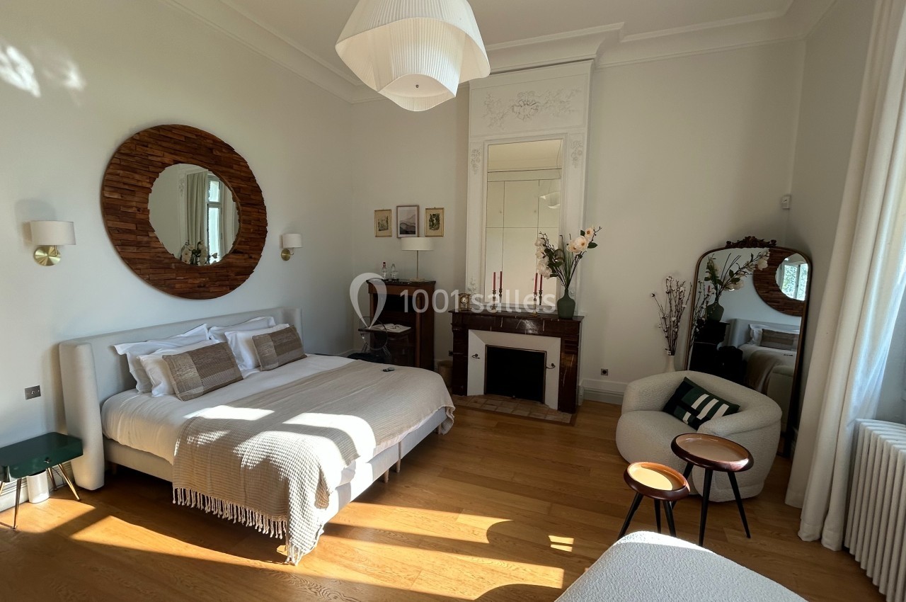 Chambre lumineuse avec lit double, miroir rond, cheminée décorative, fauteuils et parquet en bois clair.