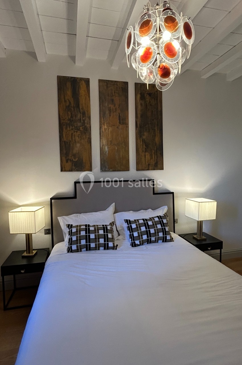 Lit double avec coussins à motifs, deux lampes de chevet allumées et un lustre moderne sous un plafond blanc.