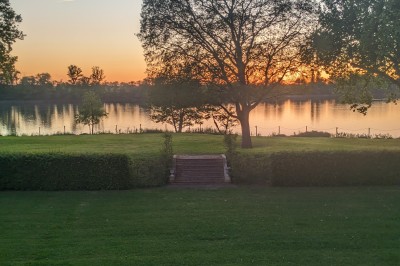 Vue d'un jardin verdoyant avec un arbre central, un escalier en pierre et un lac au coucher du soleil.