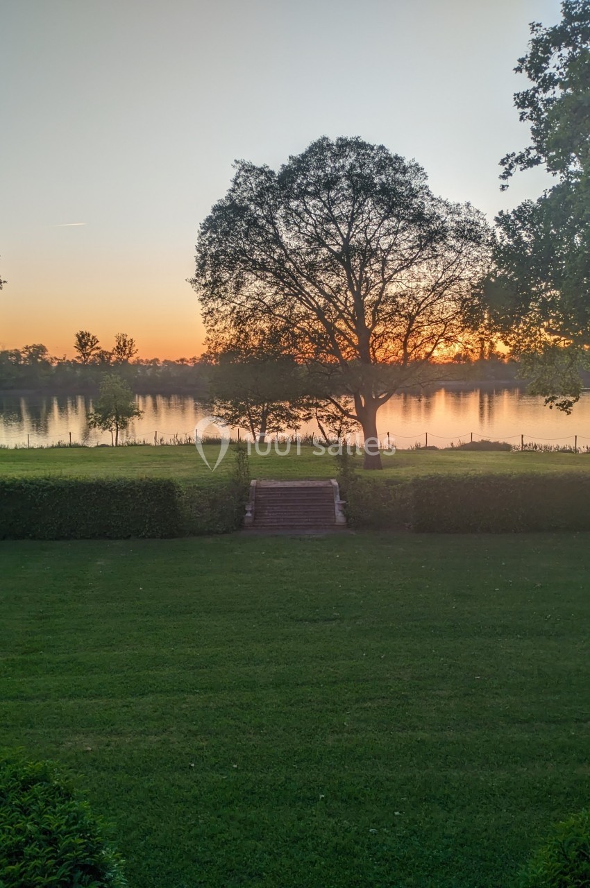 Vue d'un jardin verdoyant avec un arbre central, un escalier en pierre et un lac au coucher du soleil.