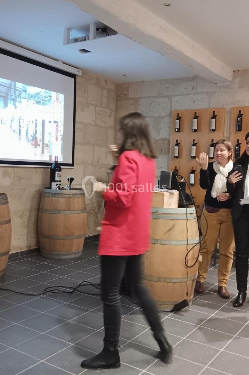 Une femme en rouge marche vers un écran de projection dans une salle avec des tonneaux et des bouteilles de vin exposées.