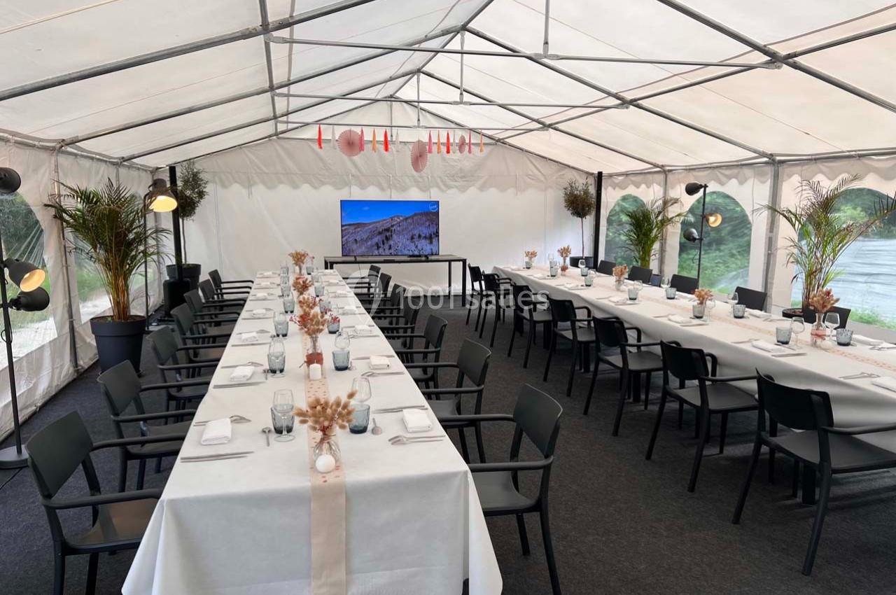 Salle de réception sous tente avec tables dressées, chaises noires, décorations florales et écran en fond.