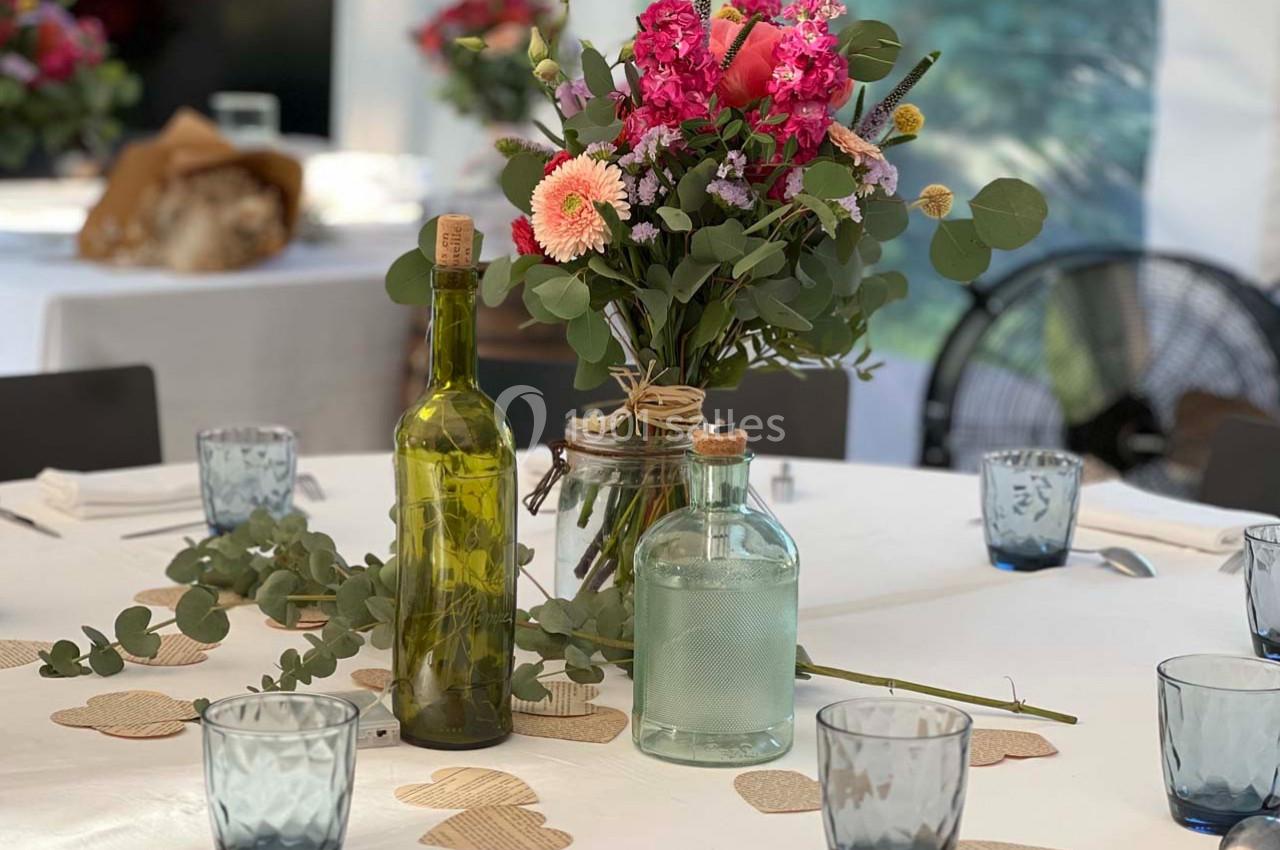 Centre de table avec bouquet de fleurs colorées, bouteilles en verre et décorations en papier sur une nappe blanche.