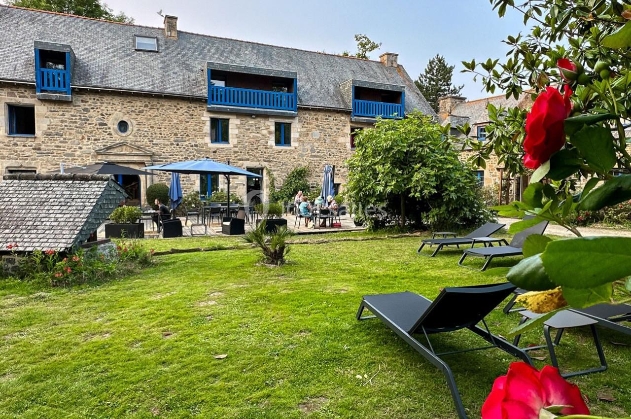 Jardin avec pelouse, chaises longues, terrasse aménagée et façade en pierre d'une maison avec balcons bleus.