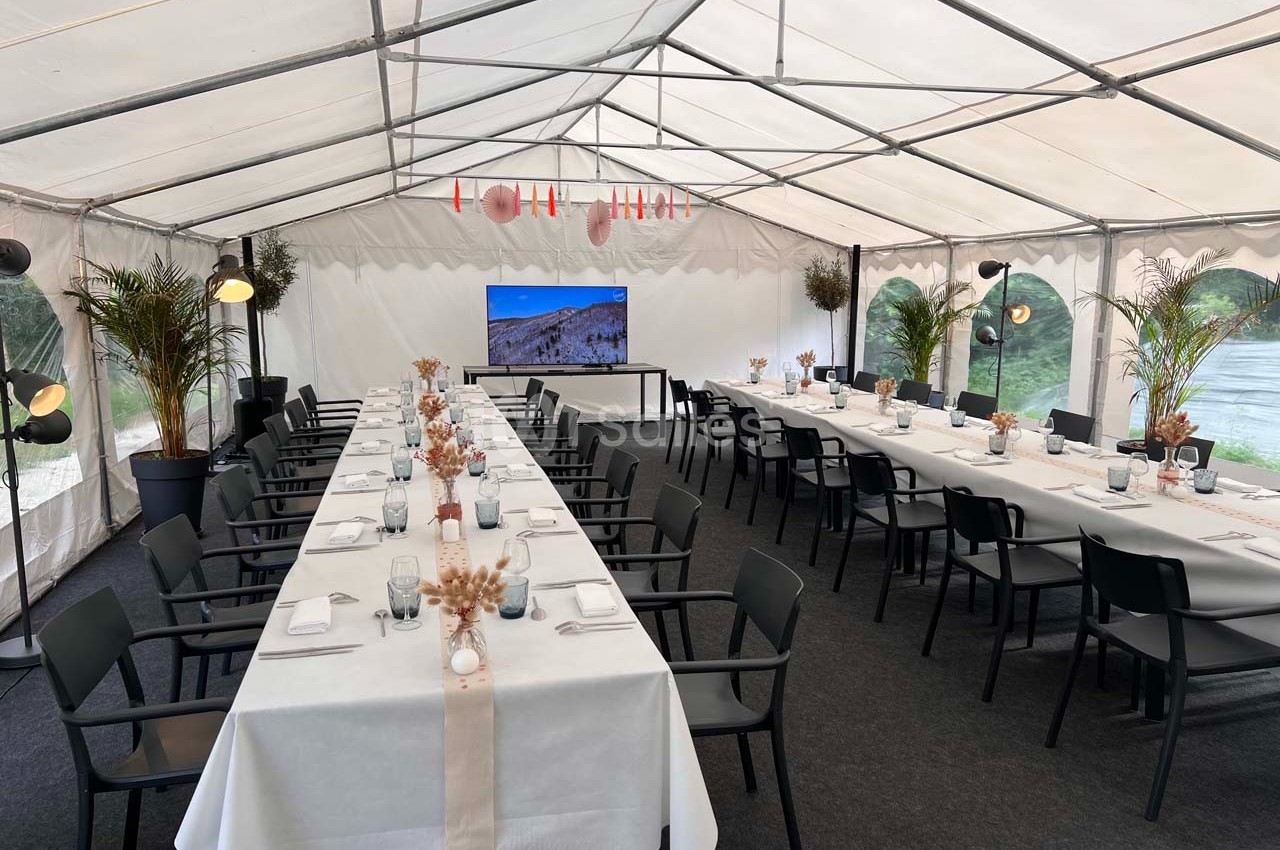 Salle de réception sous tente avec tables dressées, chaises noires, décorations florales et écran en fond.