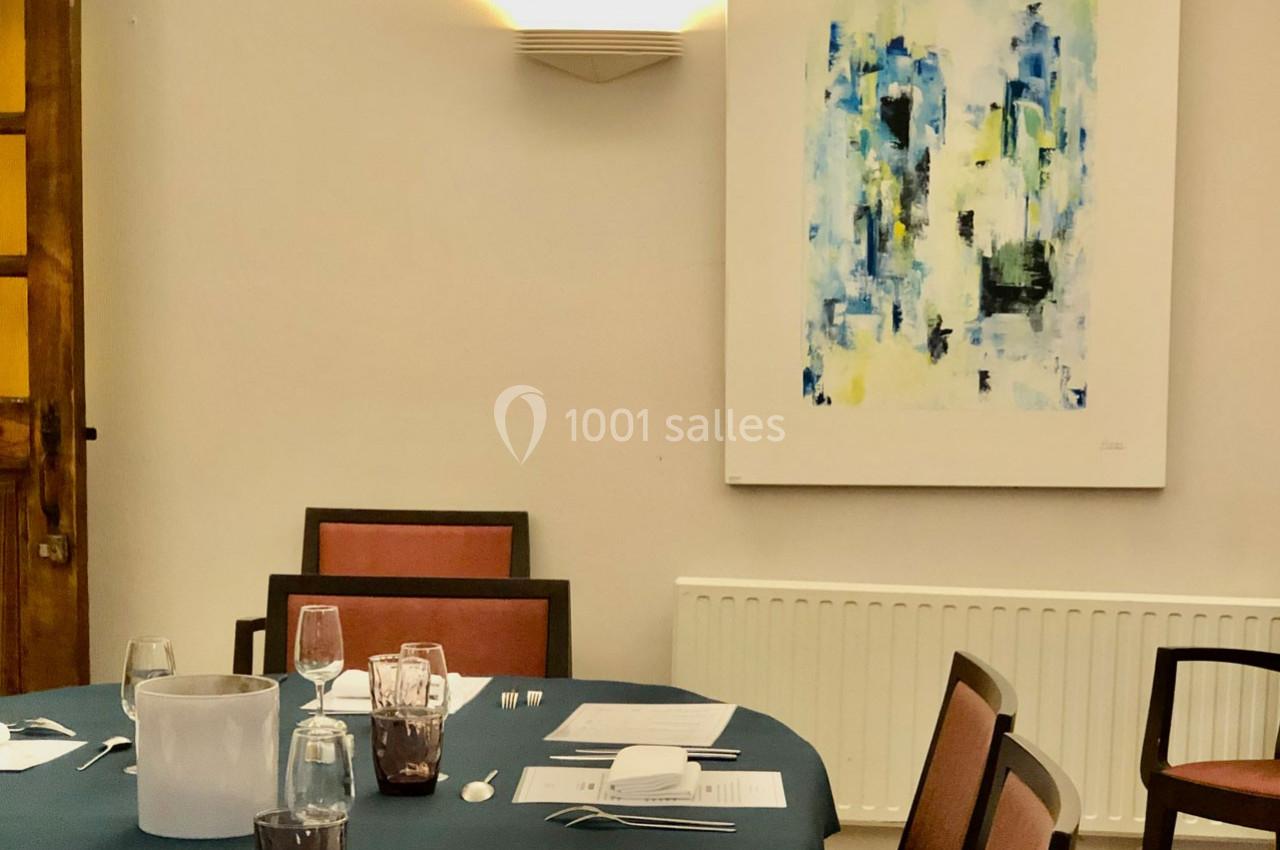 Table dressée avec nappes bleues, verres et couverts, entourée de chaises, dans une salle avec un tableau abstrait au mur.