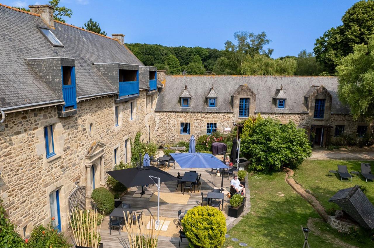 Cour intérieure d'un bâtiment en pierre avec terrasse aménagée, parasols, chaises longues et végétation environnante.