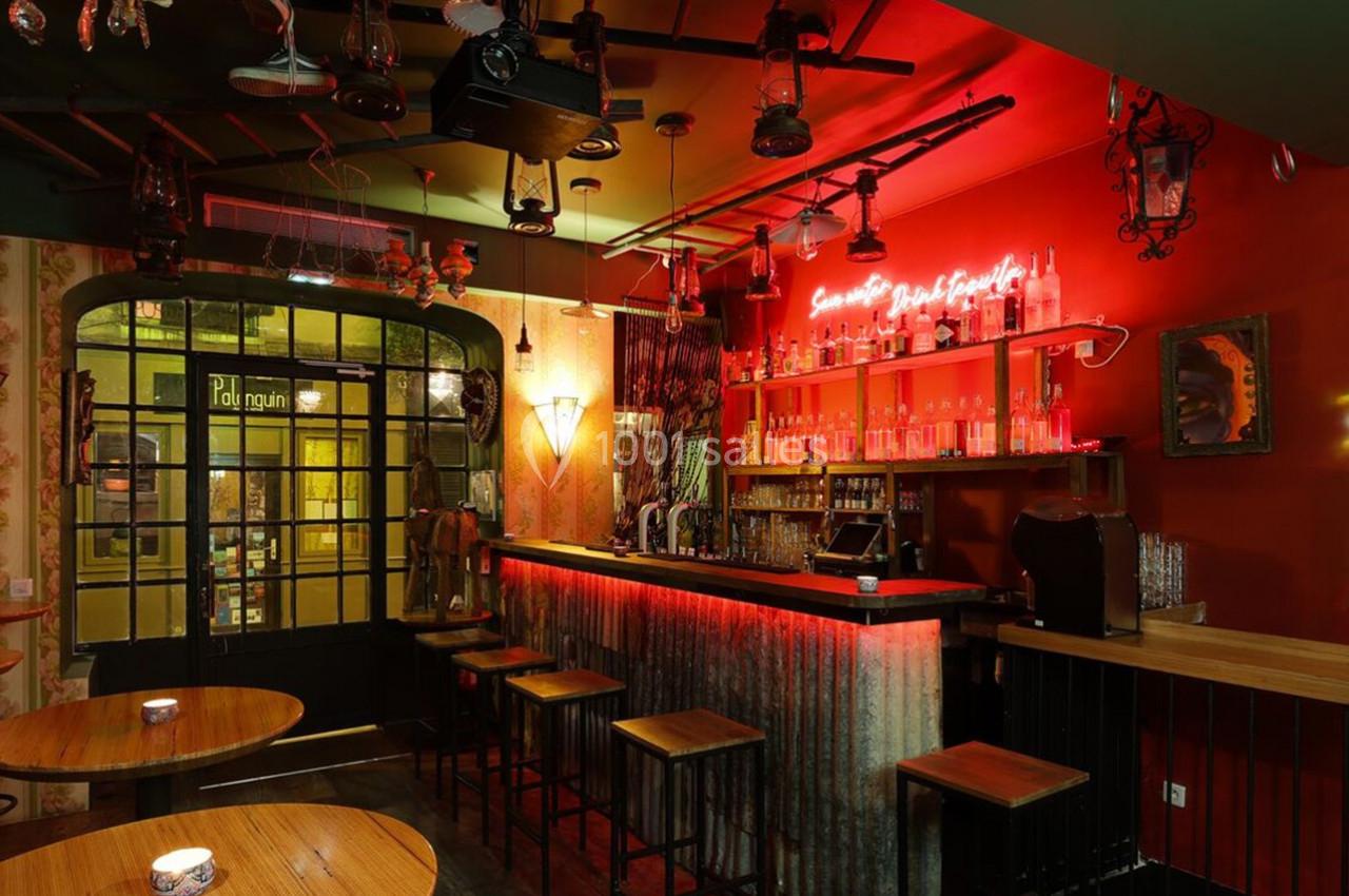 Bar à l'ambiance chaleureuse avec éclairage tamisé, comptoir en métal, tabourets hauts et décorations murales éclectiques.