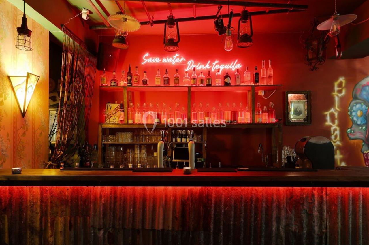 Bar avec éclairage rouge, étagères remplies de bouteilles, néon au mur et décoration colorée sur la droite.