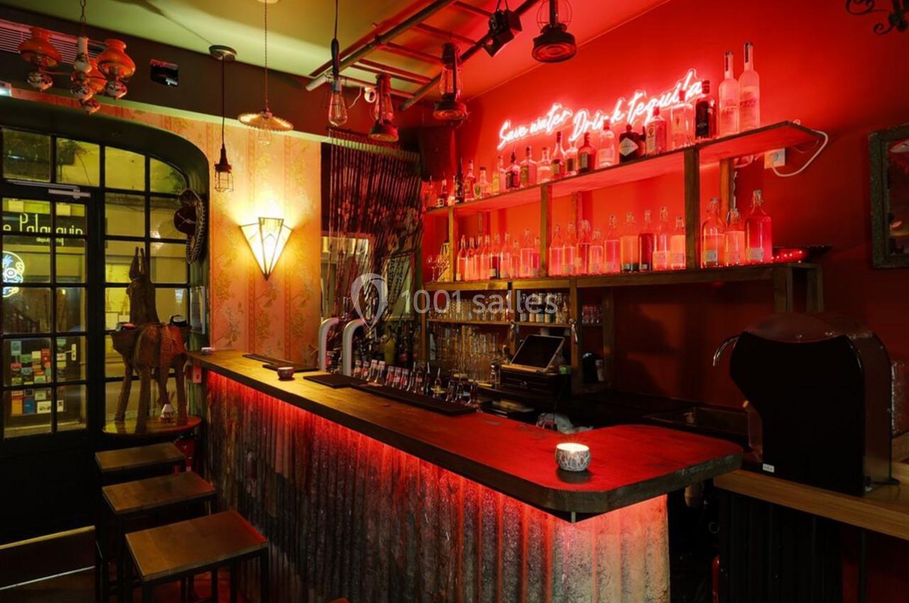 Bar à l'ambiance chaleureuse avec éclairage rouge, comptoir en bois, étagères de bouteilles et enseigne néon.