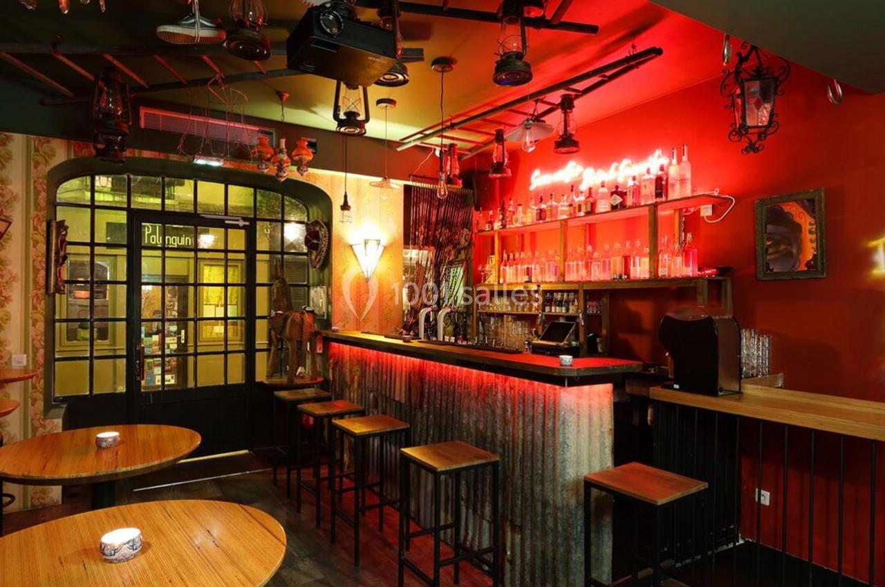 Intérieur d'un bar avec comptoir en métal, tabourets hauts, éclairage néon rouge et décorations murales éclectiques.