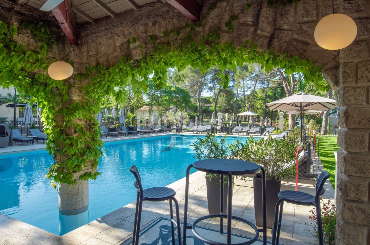 Terrasse ombragée avec table et chaises donnant sur une piscine entourée de transats et de végétation.