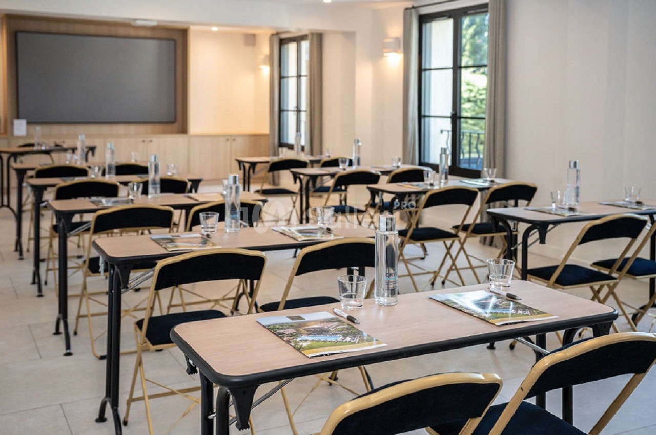 Salle de réunion lumineuse avec des tables alignées, des chaises pliantes et des documents posés sur chaque table.
