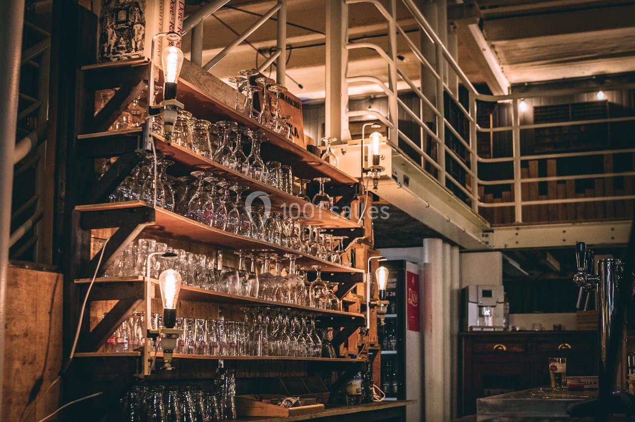 Étagères en bois remplies de verres dans un bar à l'ambiance industrielle avec éclairage tamisé.
